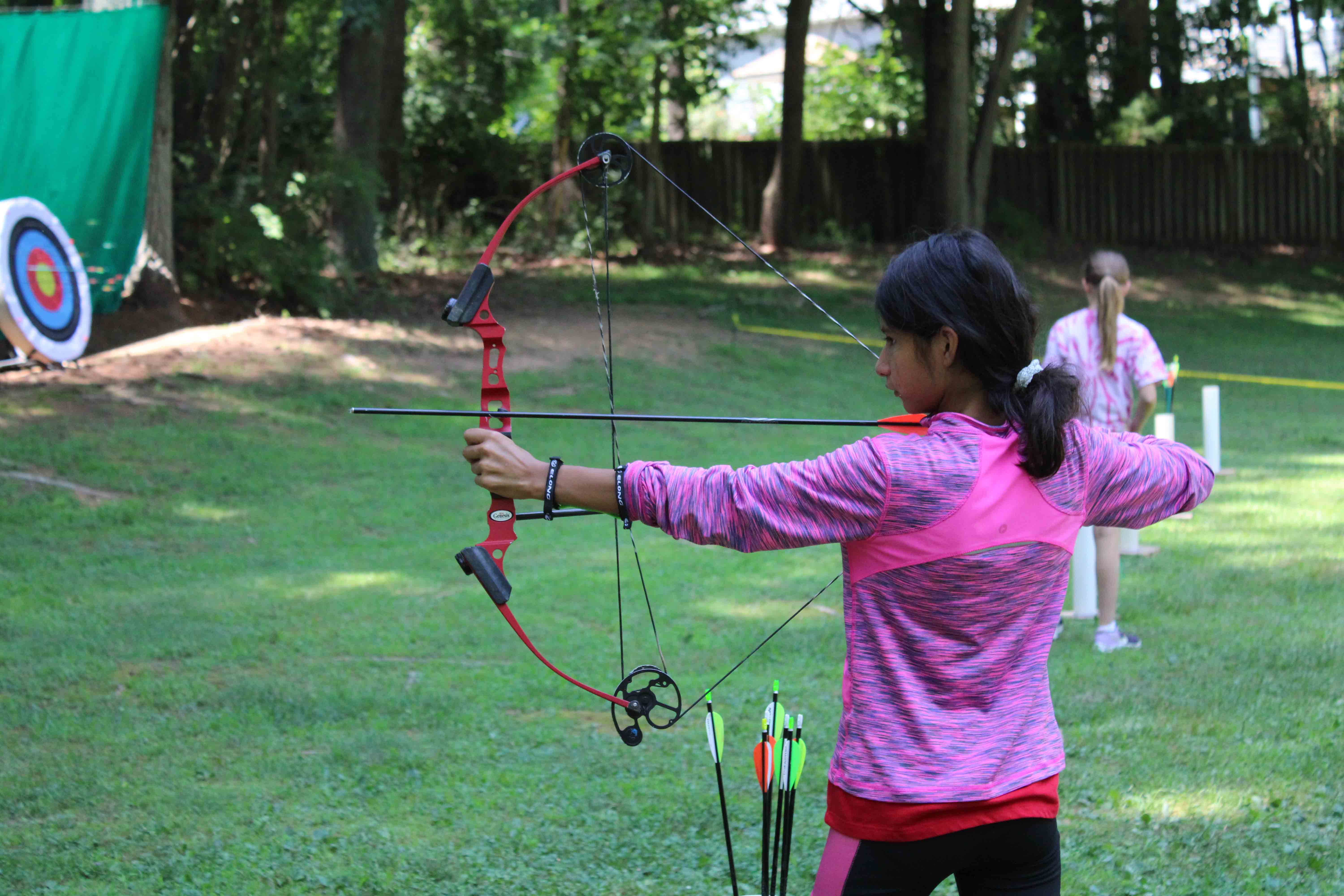 A camper from the Catapults unit wearing a pink shirt pulling back a compound bow string at the archery range.