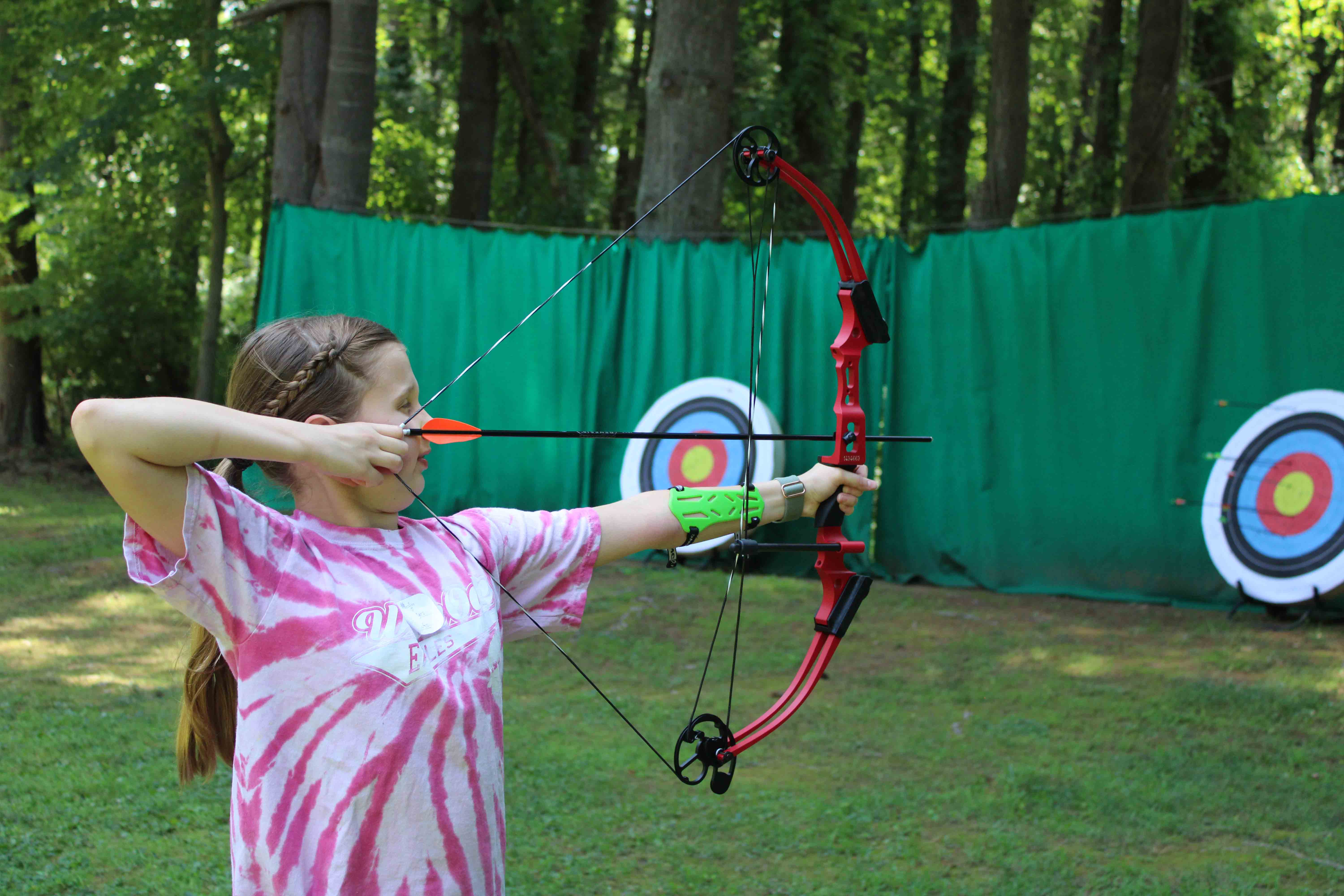 A girl in a tie-dyed t-shirt carefully aiming her bow and arrow toward a target down at the camp archery field.