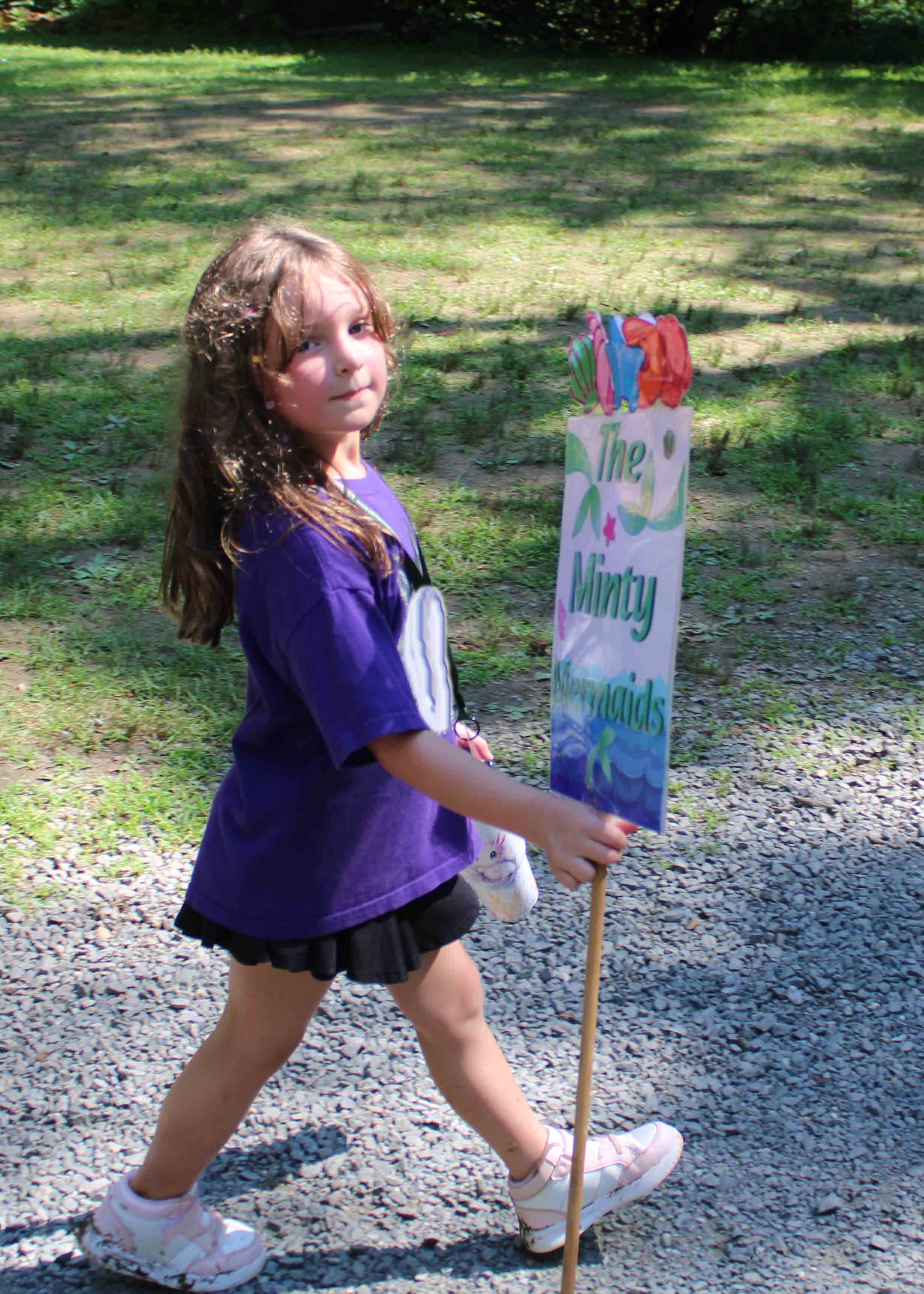 A happy camper proudly holding up her purple Minty unit banner outdoors.